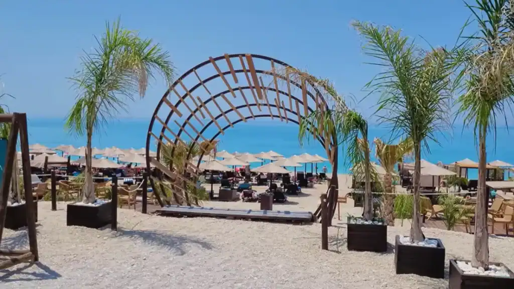 A curved wooden lattice arch flanked by potted palms leads onto a beach club's straw-umbrella terrace, turquoise water filling the gap beyond rows of sunbeds
