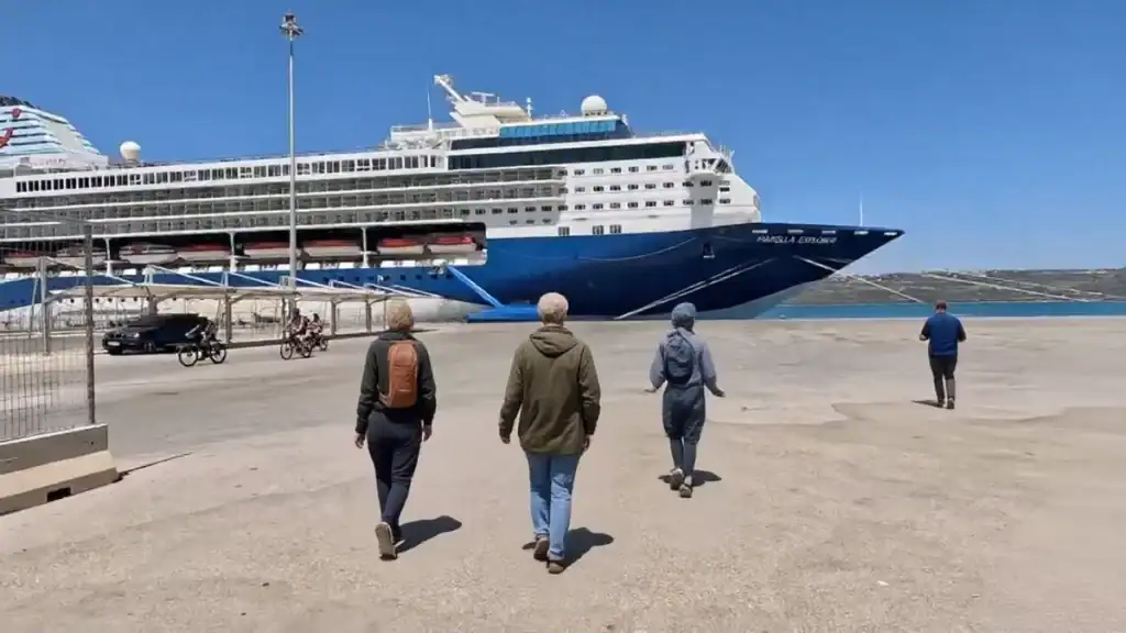 Passengers walking across Souda dock toward the Marella Explorer cruise ship berthed at the terminal, a second vessel partially visible to the left