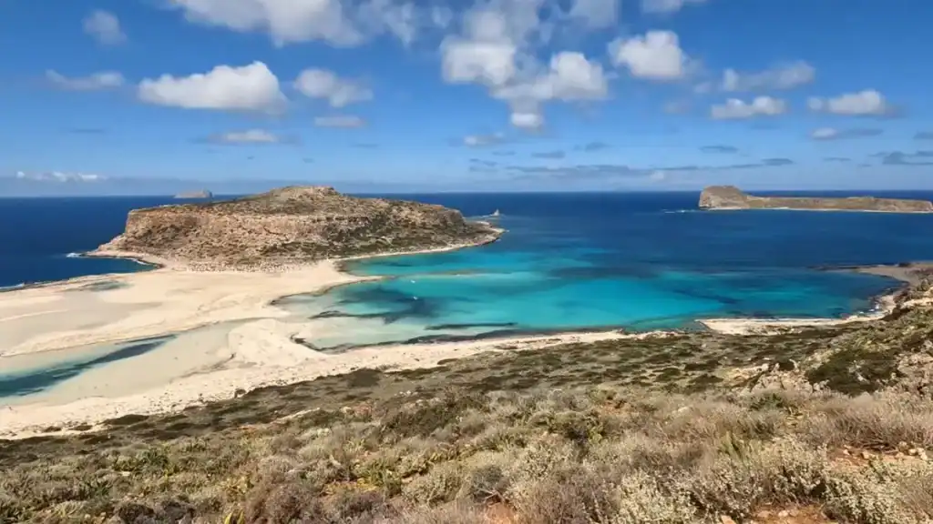 Balos lagoon seen from the ridge above, its pale sandbar curving between vivid turquoise shallows and deep blue sea, not far from Falasarna on Crete's northwest tip.