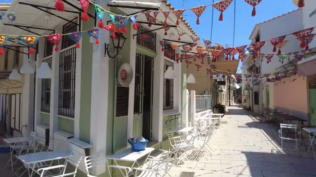 Crocheted bunting strung across a narrow alley in Lefkada town, where pastel-fronted café buildings line a stone-paved lane set with empty white bistro chairs.