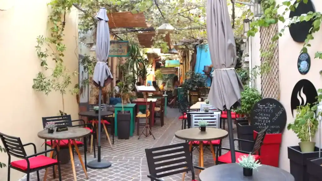 Narrow vine-covered alley café in the old town, red-cushioned chairs around small round tables with folded parasols and potted plants on both sides