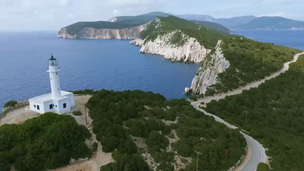 Cape Lefkatas lighthouse standing white against the Ionian Sea, a winding road cutting through dense scrub toward the sheer limestone cliffs of the headland beyond.