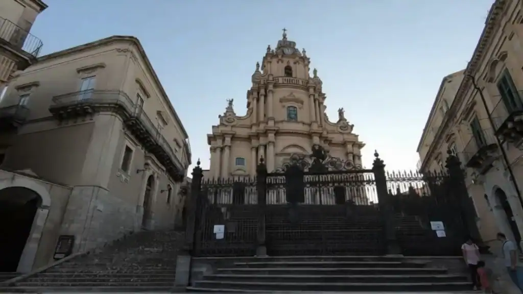 Facade of the Cathedral of San Giorgio rising above a broad flight of steps, its tiered baroque stonework framed by flanking buildings and closed iron gates