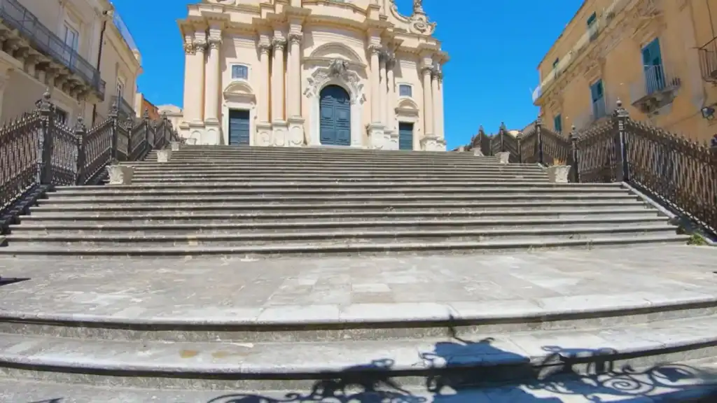 Wide stone staircase climbing to the ornate Baroque facade of the Cathedral of San Giorgio, wrought-iron railings casting shadows across the steps in the Ragusa valley hill town of Ragusa Ibla