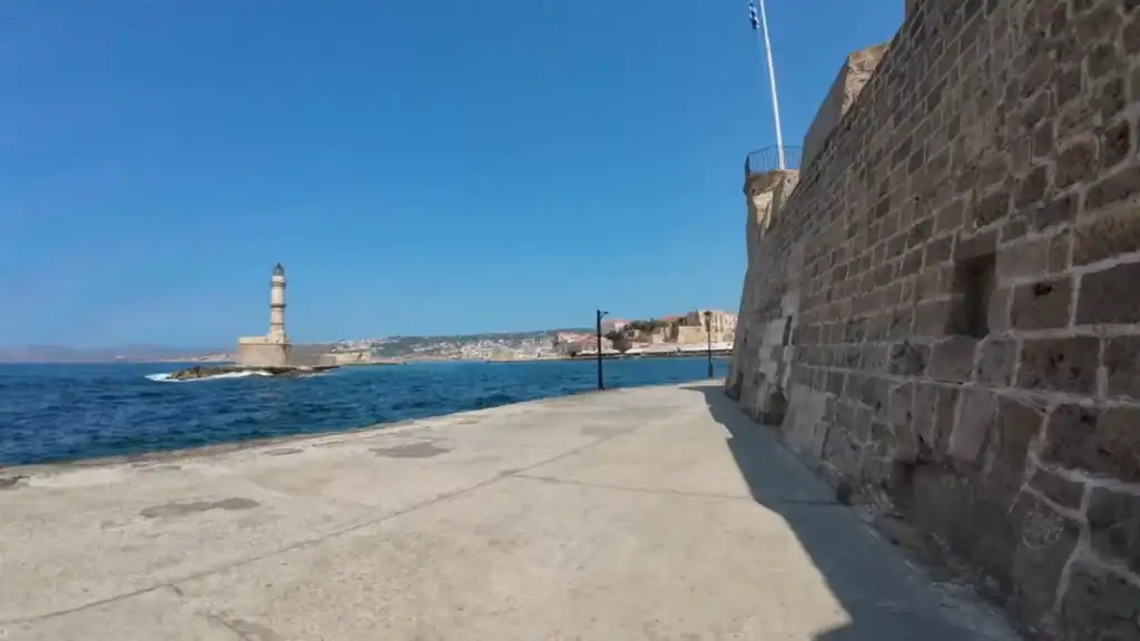 A wide harbour walkway runs alongside a thick stone fortress wall, with a lighthouse standing at the end of the breakwater under a clear blue sky
