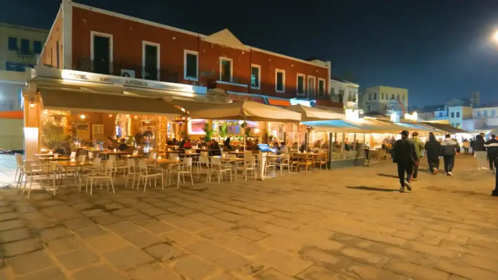 Harbour-front restaurants strung with warm fairy lights on a Chania in November evening, diners seated under awnings as pedestrians cross the wide stone-paved quay