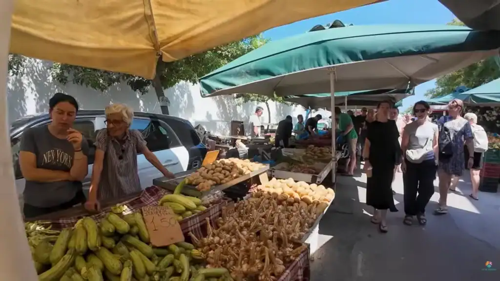Outdoor farmers market stalls piled with courgettes and root vegetables, shoppers walking in Chania under green canvas awnings