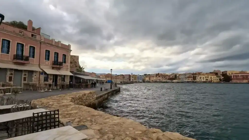 Empty cafe tables along the Chania old harbour waterfront, Venetian buildings in faded pink and ochre lining the promenade under heavy cloud cover