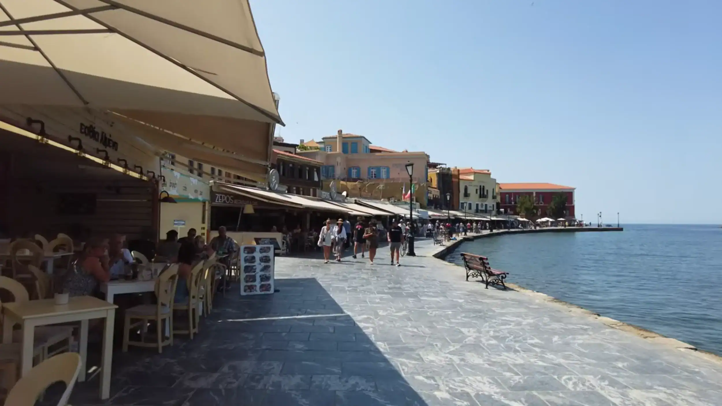 Chania's Old Harbour waterfront on a clear morning, with taverna seating along the promenade and colourful Venetian buildings curving toward the sea — an easy stretch to cover on solo travel in Chania at a relaxed pace.