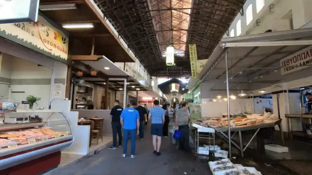 Shoppers moving through the central aisle of Chania Municipal Market, fish displayed on the right counter and a poultry stall visible on the left beneath a vaulted iron roof