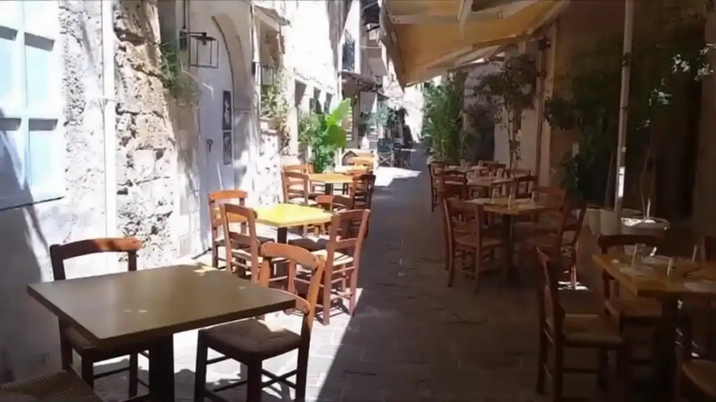 Empty wooden tables and chairs fill a sun-dappled narrow lane in Chania Old Town, potted greenery tucked between stone walls on both sides