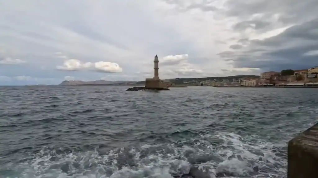 Chania lighthouse in December standing at the harbour entrance as rough seas churn beneath an overcast sky, with the old town visible along the waterfront