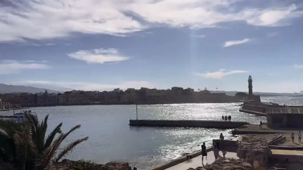 Lighthouse silhouetted on a stone breakwater across glittering harbour water, old town rooftops stretching behind under a partly clouded sky