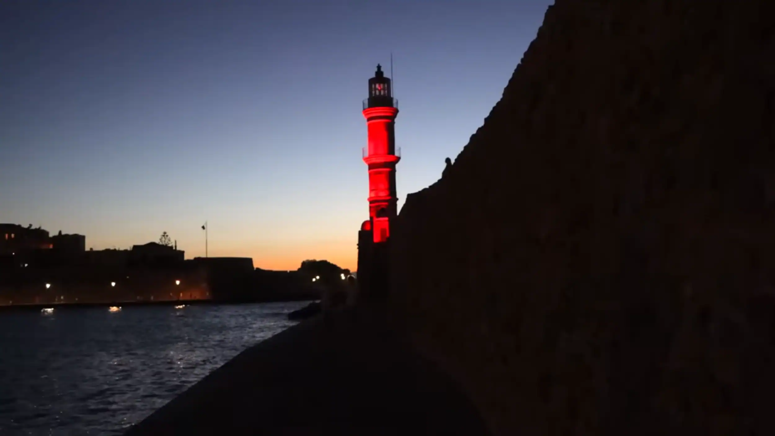 Chania lighthouse lit red against a deep blue dusk sky, seen from the darkened harbour wall