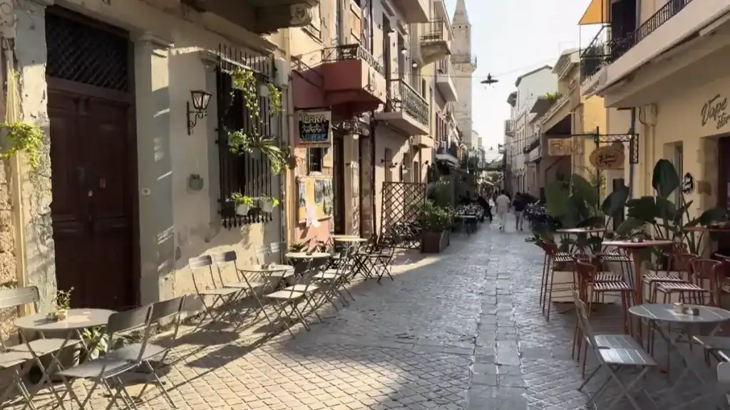 Cobbled side street with cafe tables on both sides, potted plants climbing weathered facades, a minaret visible in the distance