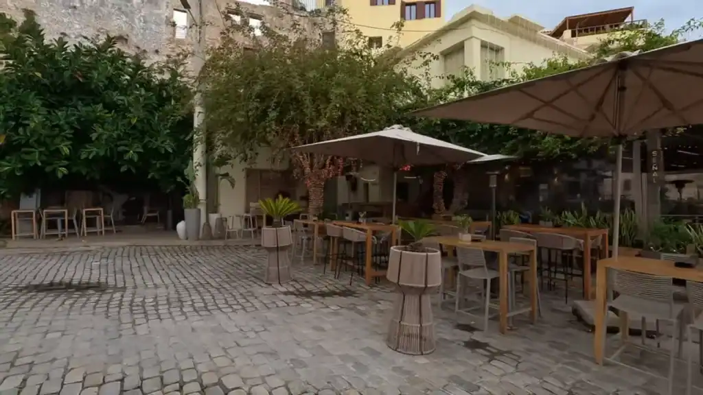 Deserted outdoor restaurant terrace on a cobbled square in Chania in December, wooden tables and parasols set but unoccupied, mature trees and old stone walls surrounding the space