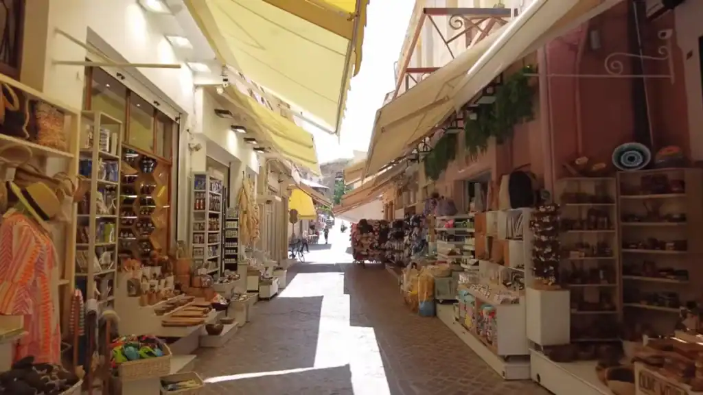 Narrow old town lane in Chania lined on both sides with open shop fronts selling ceramics, hats, textiles and souvenirs under retracted canvas awnings.