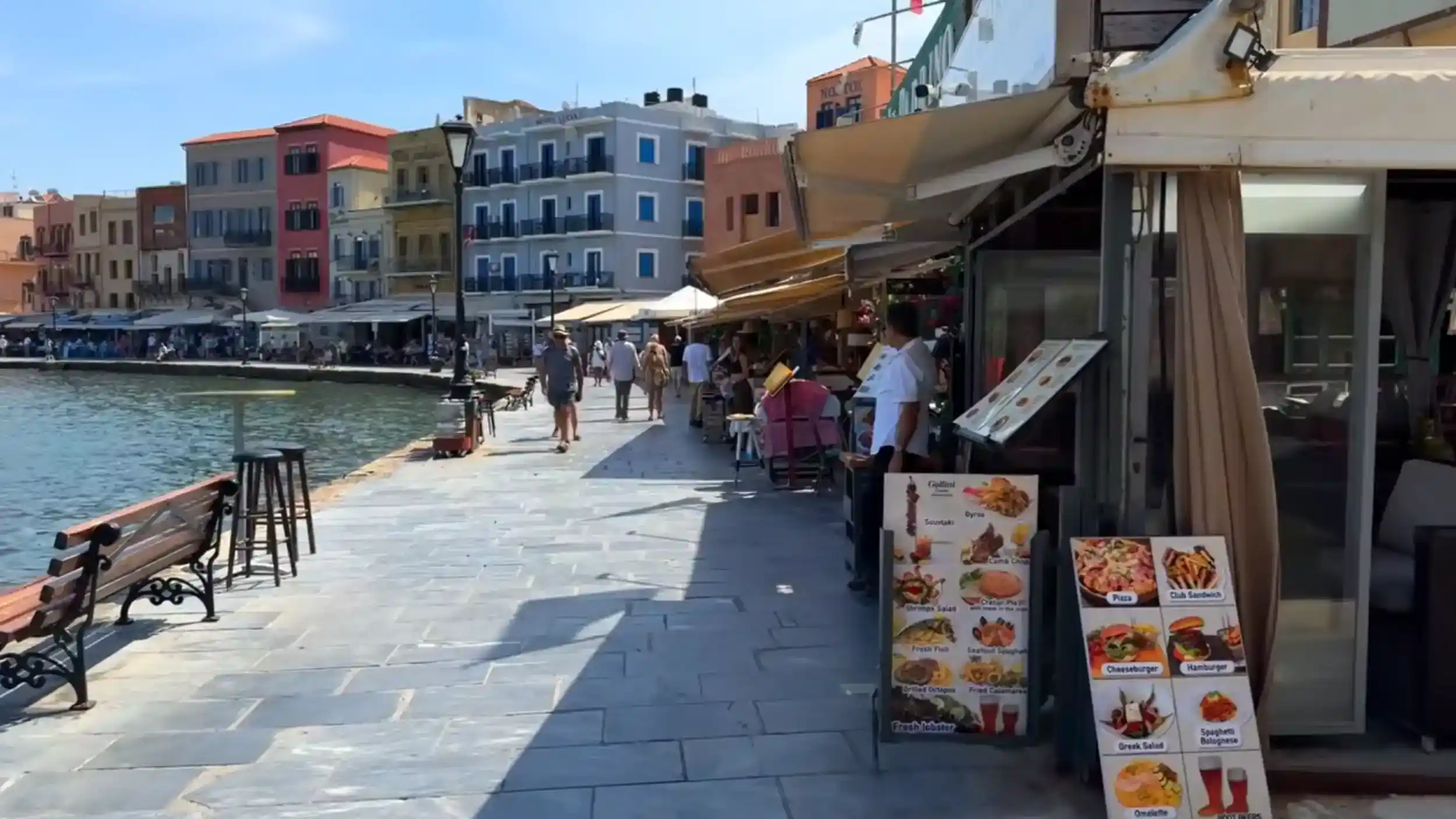 Taverna staff reviewing a menu board on the Chania old harbour waterfront, coloured buildings lining the far quay after the Souda Bay to Chania route