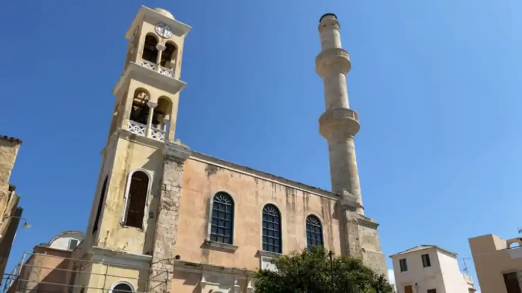 Church of Agios Nikolaos in Chania, its weathered stone facade flanked by a clock bell tower on one side and an Ottoman minaret on the other
