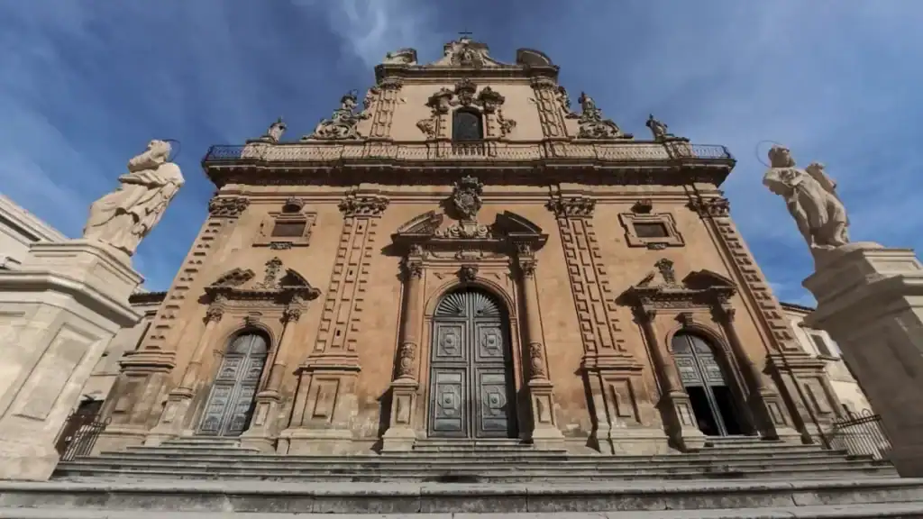Upward-angled shot of the Church of San Pietro's heavily ornamented sandstone facade, apostle statues flanking the staircase on either side, bronze doors and carved pilasters filling the frame against a partly cloudy sky.