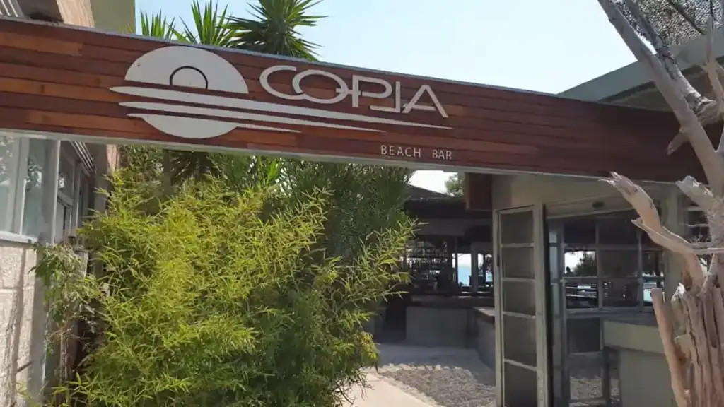 Copla Beach Bar sign mounted on a dark timber fascia above an entrance flanked by dense bamboo and a bleached driftwood trunk, the open-sided interior visible beyond