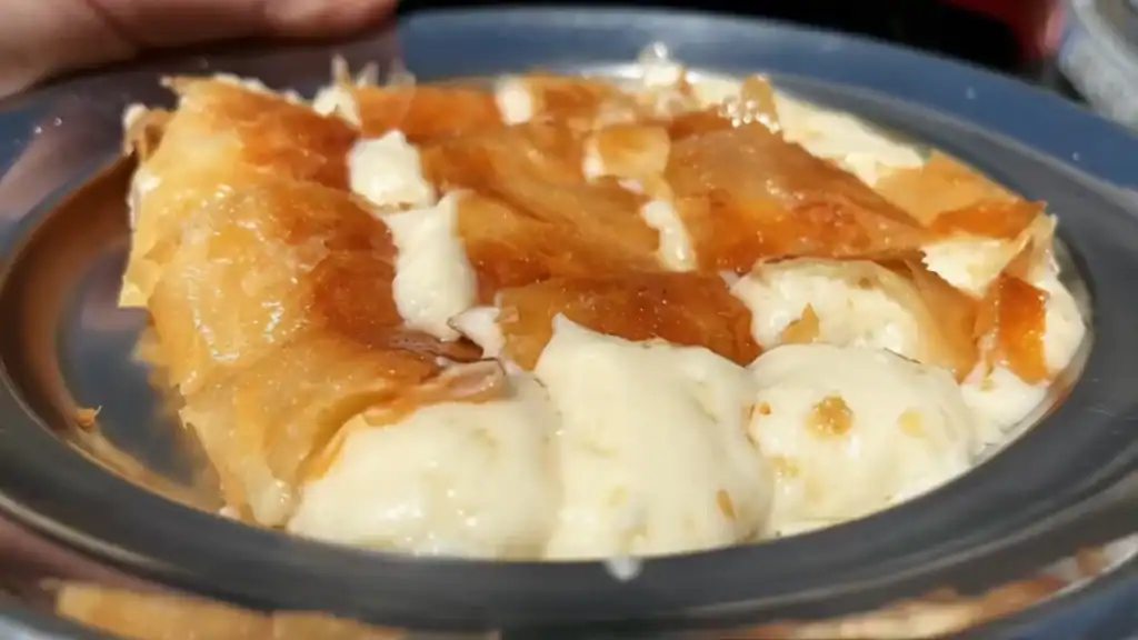 Flaky golden pastry broken open over a pool of thick cream filling on a plate, a breakfast pastry eaten fresh in Chania Old Town