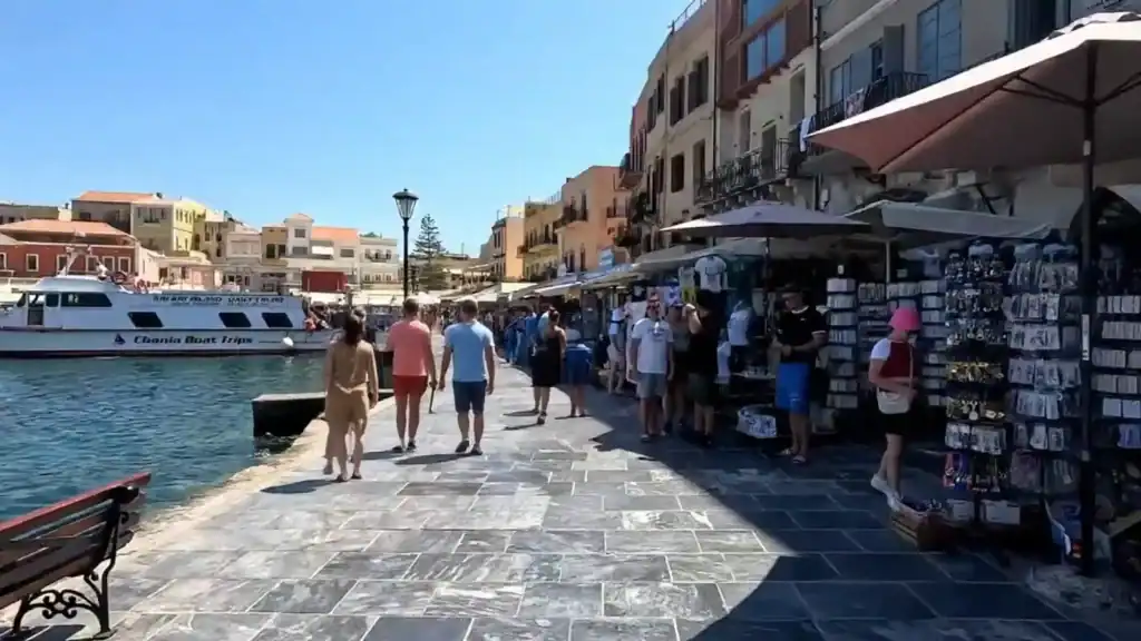 Busy harbourfront promenade in Chania's old town, what you'd typically encounter spending one day in Chania, with souvenir stalls, a moored boat trips vessel, and colourful buildings opposite
