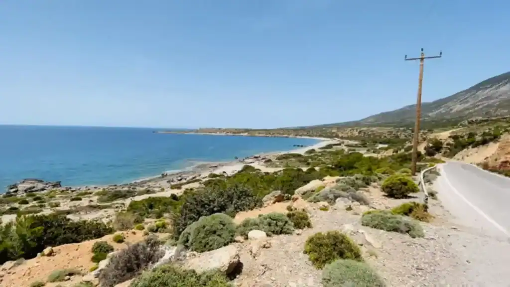 Coastal road edging past scrubland and pale rock toward Elafonisi, with a quiet sandy bay and deep blue sea stretching out to the left.