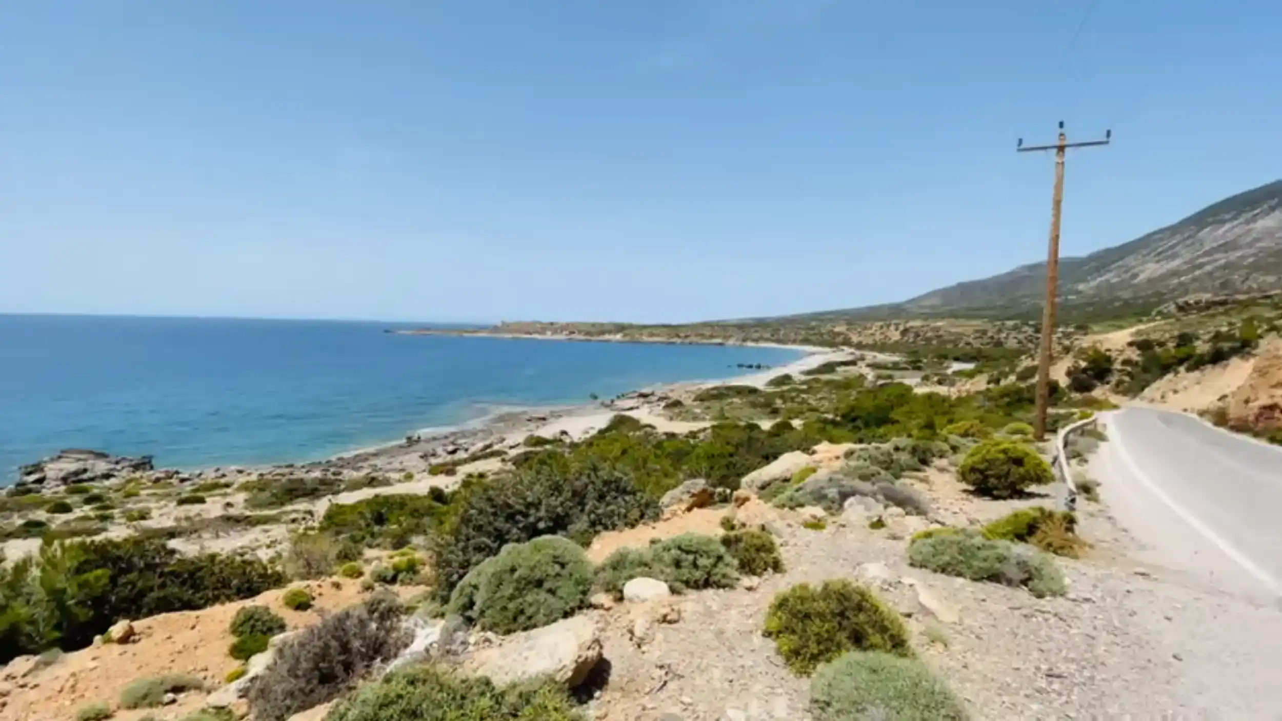 Coastal road edging past scrubland and pale rock toward Elafonisi, with a quiet sandy bay and deep blue sea stretching out to the left.