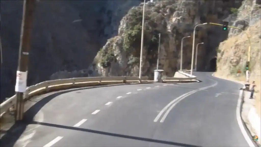 Narrow two-lane road curving toward a tunnel cut through limestone cliffs on the drive to Elafonisi, with a green traffic light overhead and metal crash barriers along the bend.