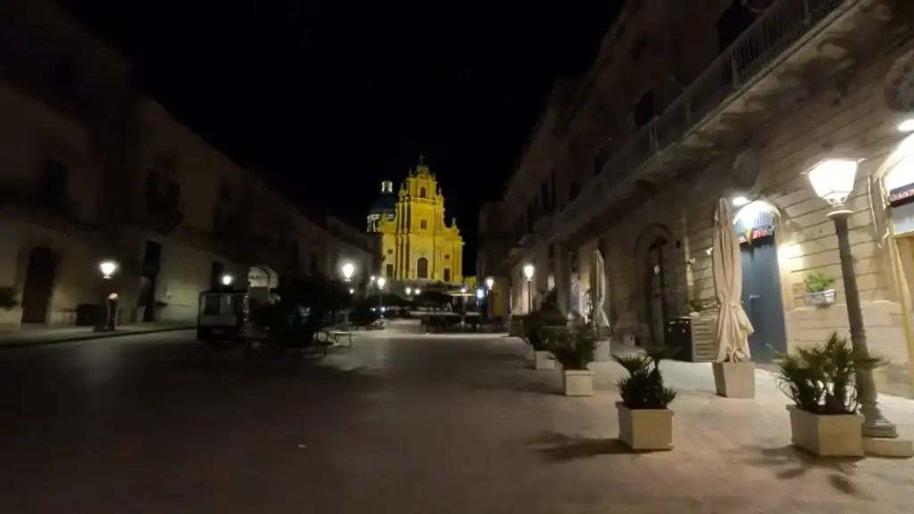 Duomo di San Giorgio floodlit in deep gold at the far end of a deserted pedestrian street, baroque façade rising against a black sky