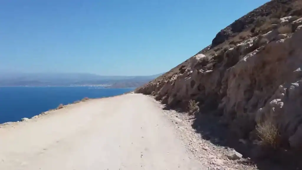 Unpaved dusty road to Balos Lagoon running along a steep rocky hillside with deep blue sea visible to the left and a distant coastline on the horizon