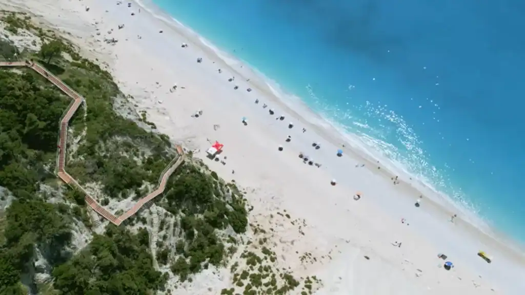 Egremni beach seen from directly above, a long strip of white sand meeting deep blue water, with a zigzagging wooden staircase descending the scrub-covered cliff on the left.