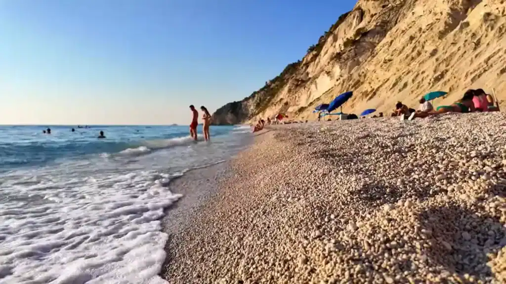 Egremni Beach shingle foreground with small waves breaking at the waterline, lefkada in may drawing swimmers and sunbathers beneath the crumbling sandstone cliffs running the length of the shore