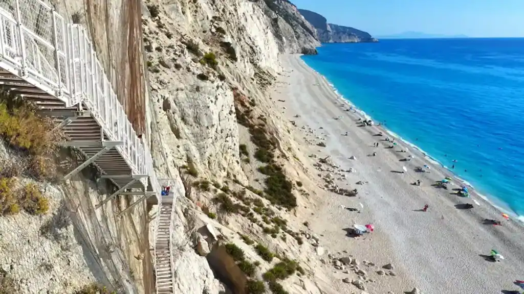 Metal staircases bolted to a sheer cliff face descending to Egremni, one of the longer Lefkada beaches stretching along the Ionian coast below