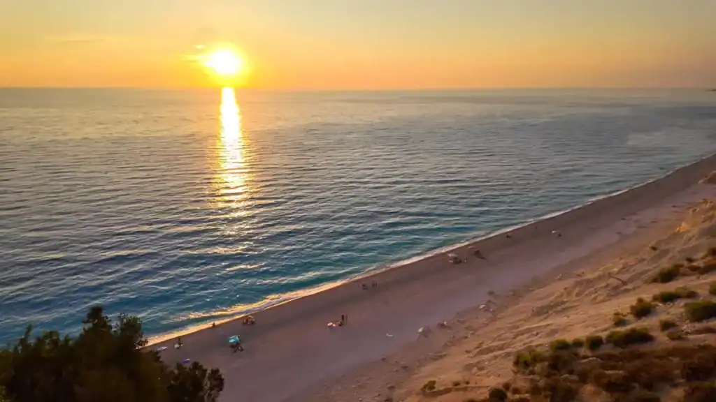 Long sandy beach running diagonally with scattered figures near the shoreline, sun low on the horizon casting a direct reflection across open sea, scrub-covered dune edge in the foreground.