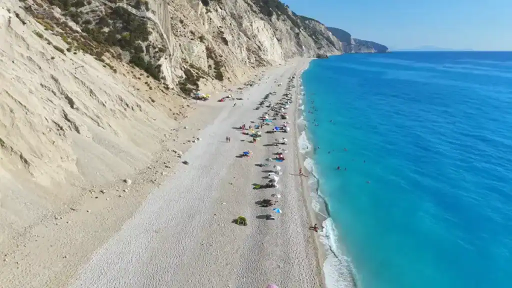 Wide pebble shore running between pale limestone cliffs and deep turquoise water, scattered with sunbathers and colourful umbrellas