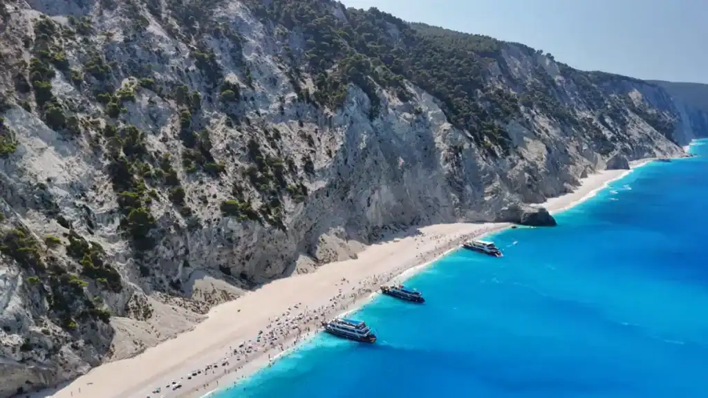 Three large tour boats moored close to a packed pebble shore, pine-covered limestone cliffs rising steeply behind and vivid blue water in the foreground