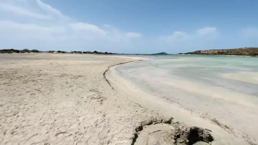 Wide flat sandbar curving along the edge of shallow turquoise water, the islet visible in the distance across the lagoon.