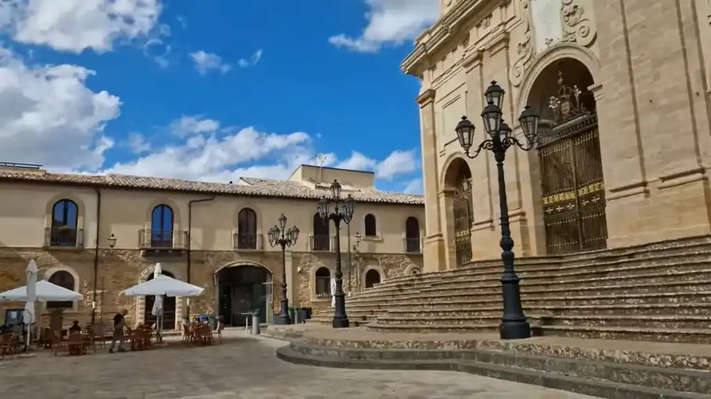 Broad stone steps and ornate facade of Enna Cathedral filling the right side of the frame, with cast-iron lamp posts and a cafe terrace spread across the piazza beside it.