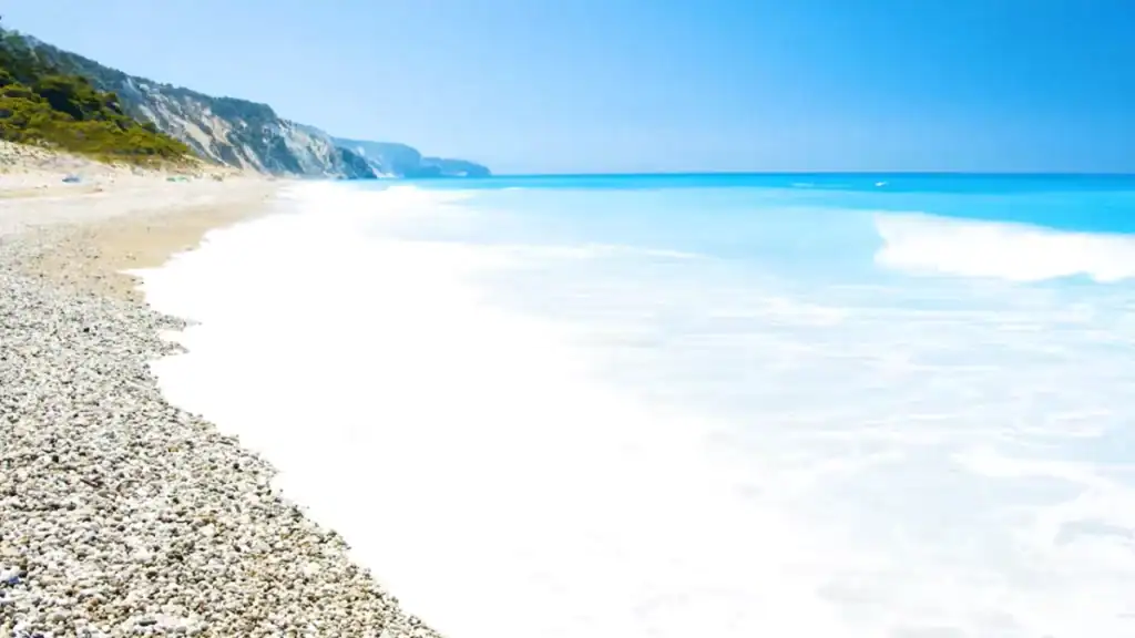Shallow surf washing over white pebbles at Porto Katsiki, the shoreline curving away toward distant cliffs, turquoise open water to the right under a clear sky.
