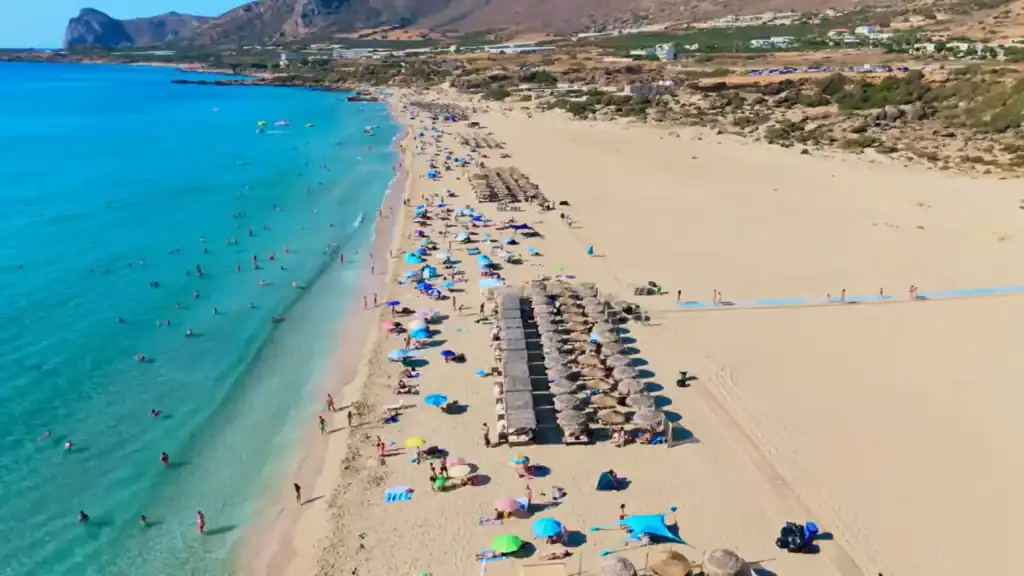 Drone shot looking down at Falasarna beach crowded with colourful umbrellas and swimmers in turquoise shallows, scrubby hillside to the right.