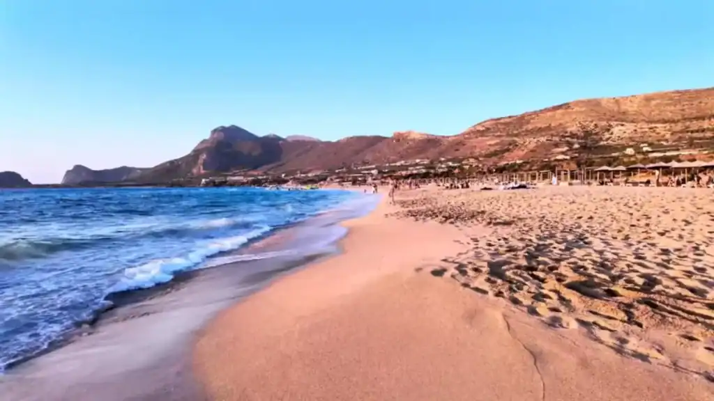 Wide sandy shore at Falasarna with shallow surf washing the beach edge, straw umbrellas lining the middle distance and dry hills rising behind.