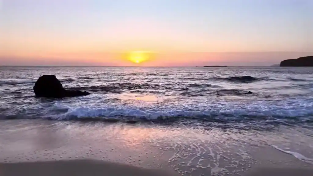 Sun touching the horizon over open sea at Falasarna, pink and amber sky reflected across wet sand, a dark rock breaking the surf in the middle ground.