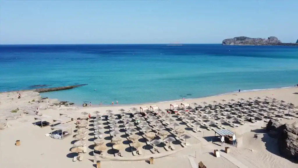 Rows of straw parasols and sunloungers filling a wide pale sand beach, with vivid turquoise water stretching to a rocky islet on the horizon