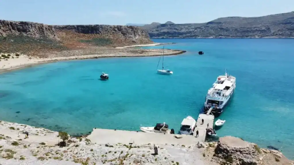 Large white ferry docked at a small concrete jetty on vivid turquoise water, with a sailboat and smaller vessels anchored nearby and a curved sandbar stretching into the bay