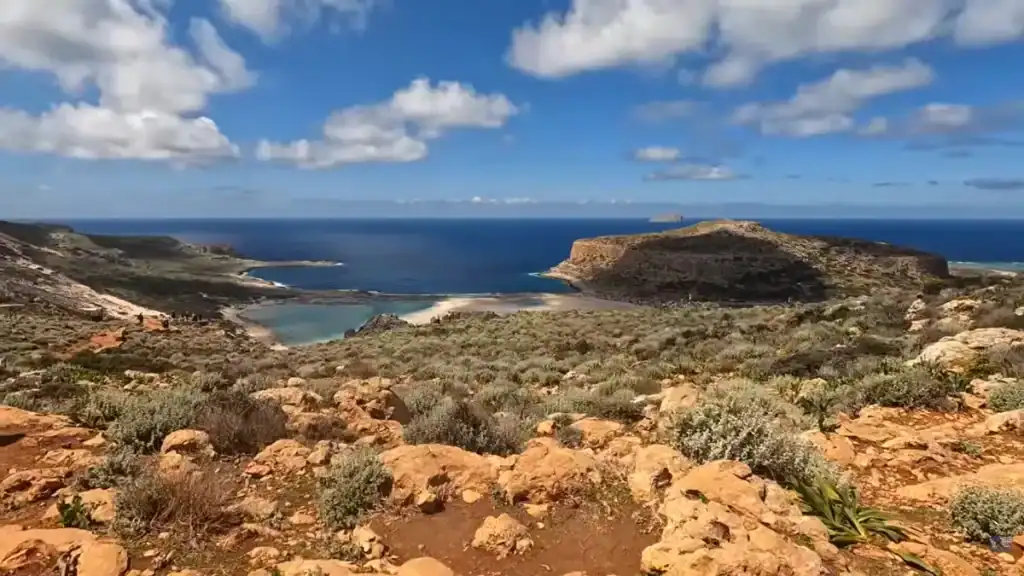 Rocky scrubland descending toward Balos Lagoon, its pale sandbar and turquoise shallows visible below a flat-topped headland with open sea and clouds beyond