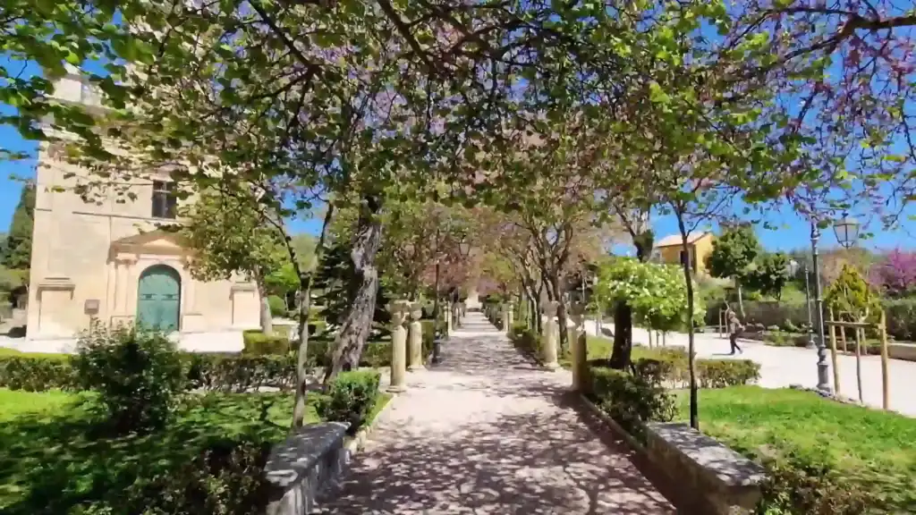 Tree-lined central path through Giardino Ibleo, branches in early blossom forming a canopy over stone column posts, a small church visible to the left
