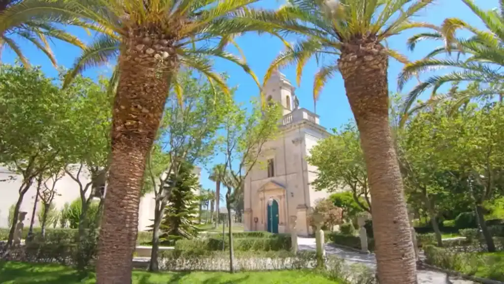 Two tall palm trunks frame a small limestone church with a square bell tower inside Giardino Ibleo, Ragusa Ibla