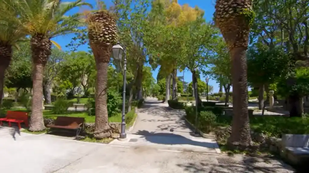 Tree-lined path through Giardino Ibleo with mature palms, iron lampposts and benches on either side, the walkway stretching into shade ahead